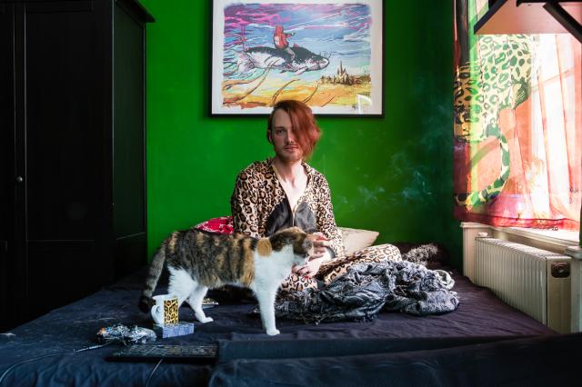 A person wearing clothes with leopard print sits on a bed. there is cigarette smoke on the air. A cat is standing also on the bed in front of them.