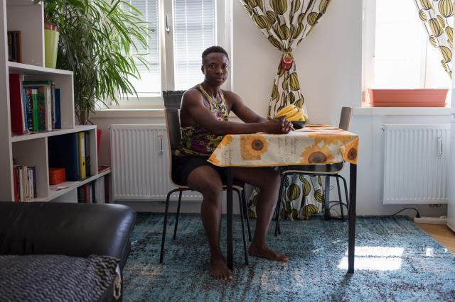 Person is sitting at a dinning room table with two chairs. There are bananas on the table. On the background a window with curtains of African green textile.