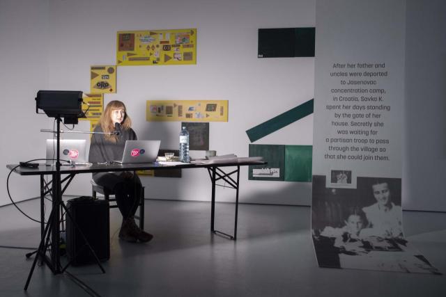 Artist sitting on a desk, talking to a public with posters having on the wall