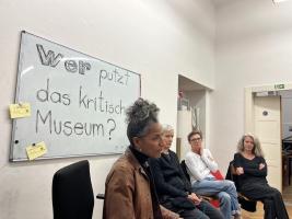 one person on the front speaking and three other people on the back sitting in a round. On the background is written on a whiteboard "Were putzt das kritische Museum"
